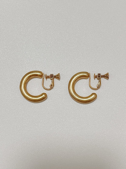 Side-by-side view of Zora solid-body brushed gold clip-on hoop earrings showing the custom matte finish and curved silhouette against a fine textured backdrop.