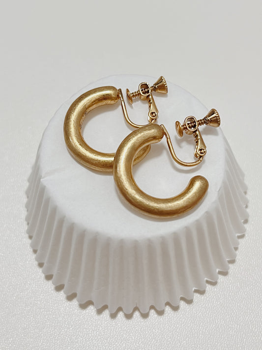 A pair of Zora matte gold clip-on hoop earrings stacked together on a white cupcake liner, highlighting the premium adjustable screw-hinge backing.