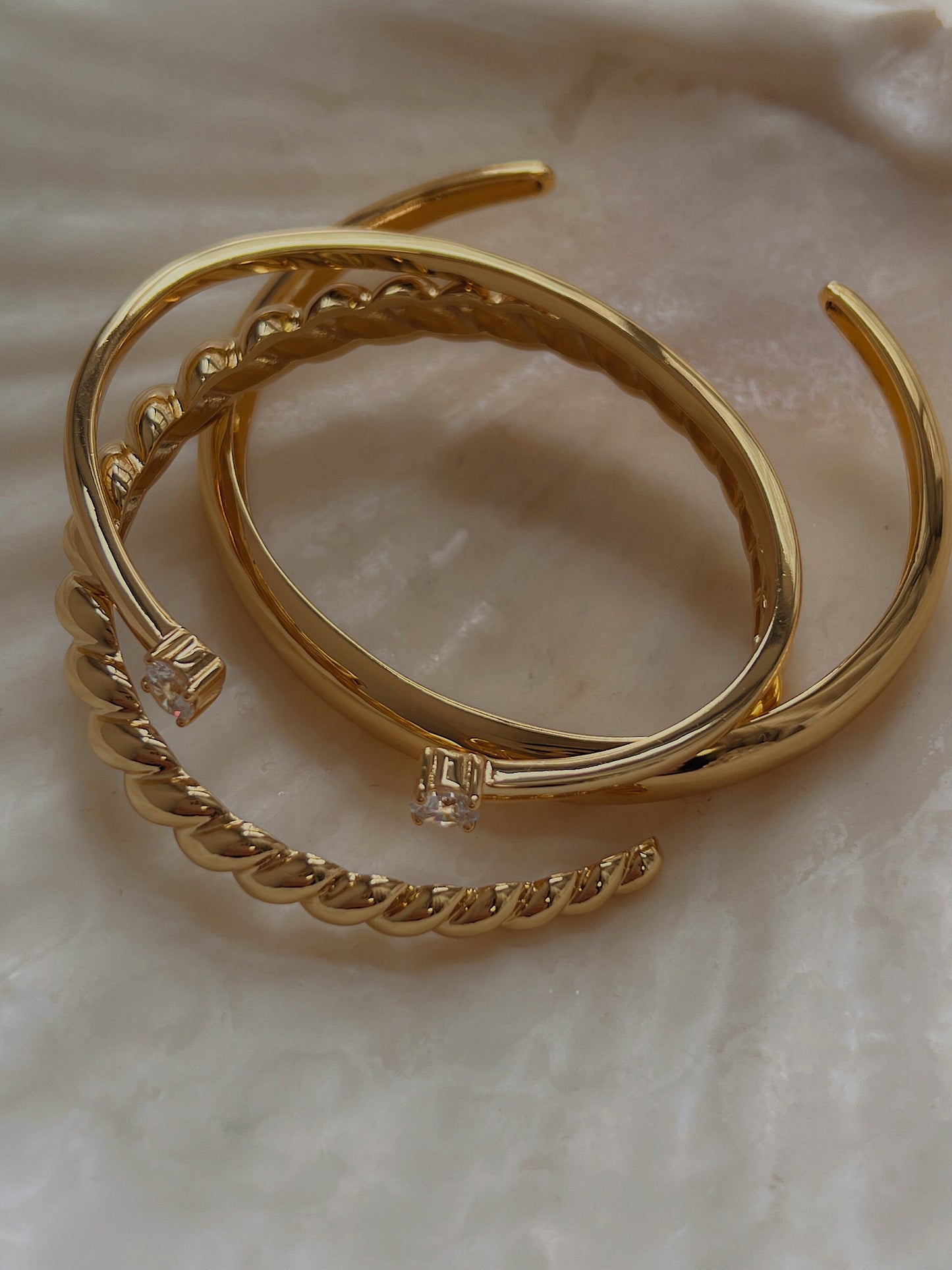 Justine gold duo-stone cuff bangles shown stacked together with a textured gold twist bangle and a minimalist smooth bangle in a shell tray, highlighting the sleek high-polish finish.
