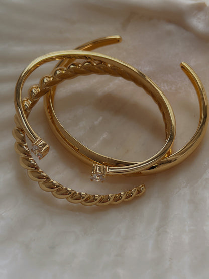 Justine gold duo-stone cuff bangles shown stacked together with a textured gold twist bangle and a minimalist smooth bangle in a shell tray, highlighting the sleek high-polish finish.