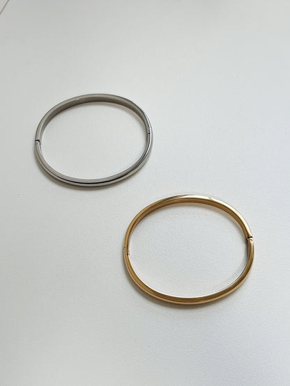 Pair of Jayden 4mm oval curved bangles in polished 18K gold and silver lying flat on a minimalist white surface.