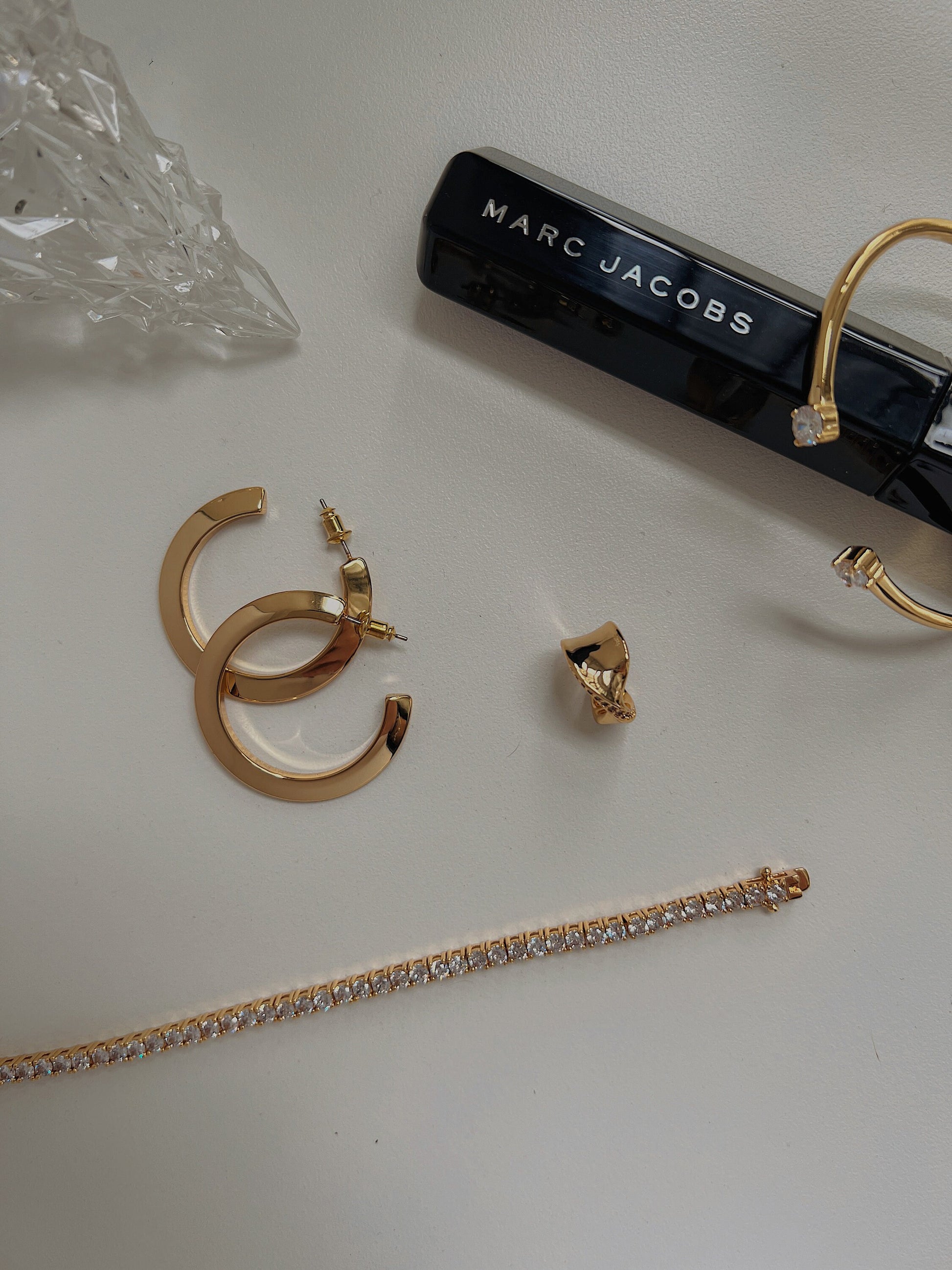 Flatlay view of the Justine gold open-cuff bracelet styled next to a crystal tennis bracelet, gold hoop earrings, and a Marc Jacobs mascara.