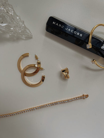 Flatlay view of the Justine gold open-cuff bracelet styled next to a crystal tennis bracelet, gold hoop earrings, and a Marc Jacobs mascara.