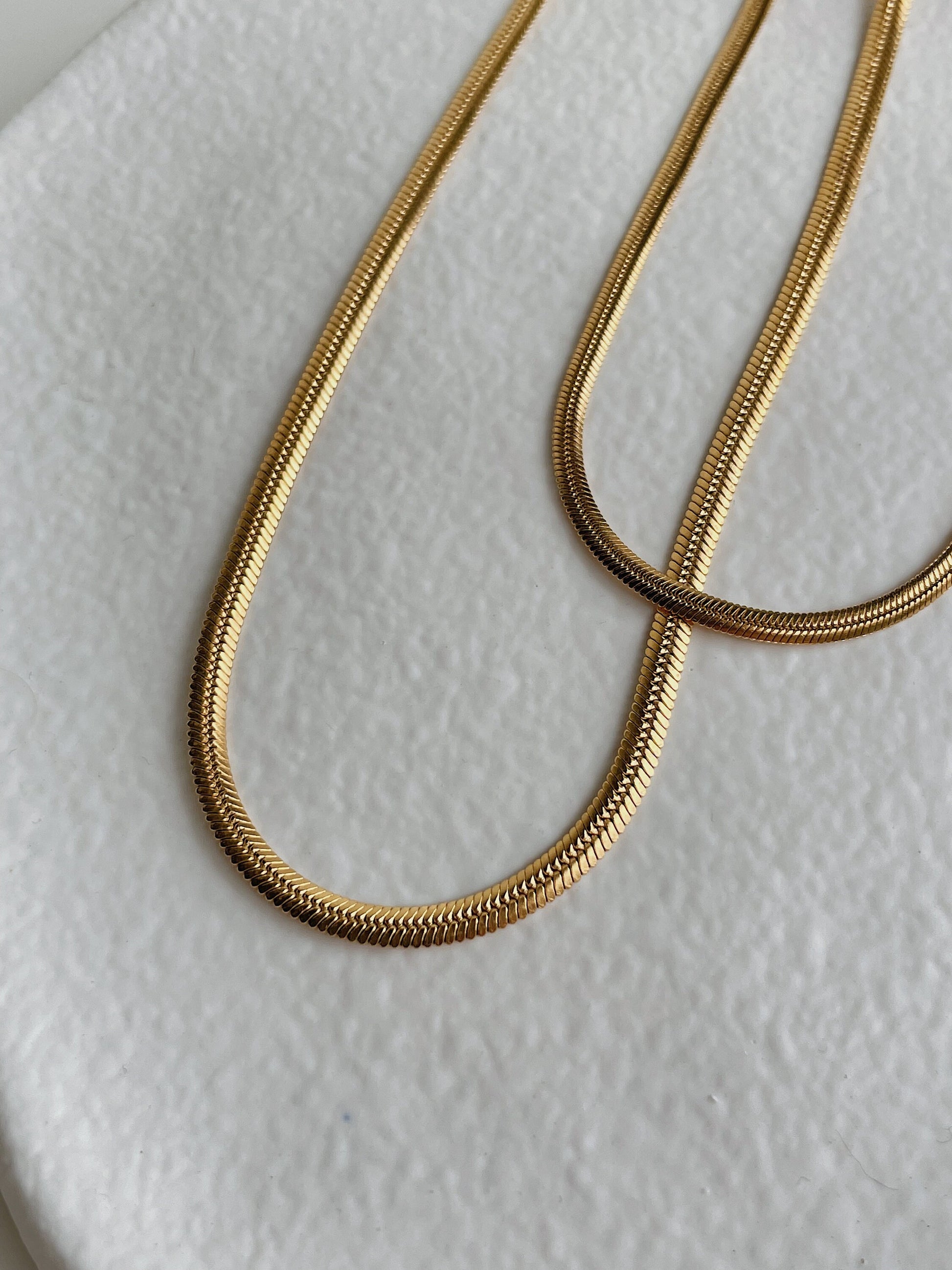 Close-up detail of 18K gold herringbone chain weave texture, warm gold tone, brass base with PVD finish
