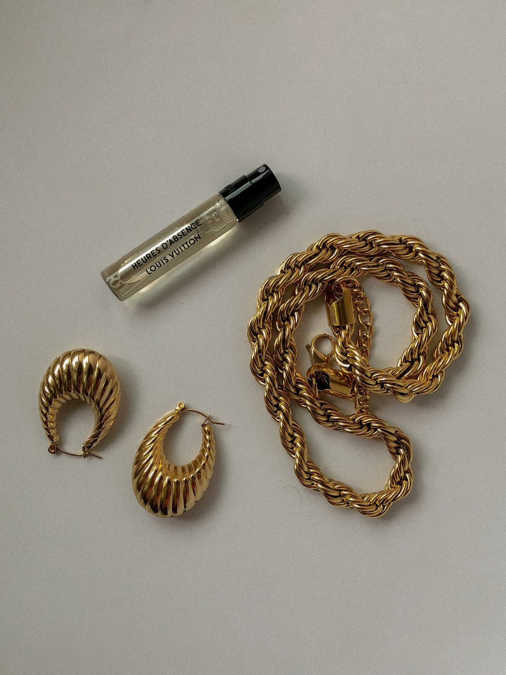 A curated luxury vanity flat lay arrangement featuring chunky ribbed gold hoop earrings, a coiled heavy twisted rope chain, and a minimalist luxury perfume vial